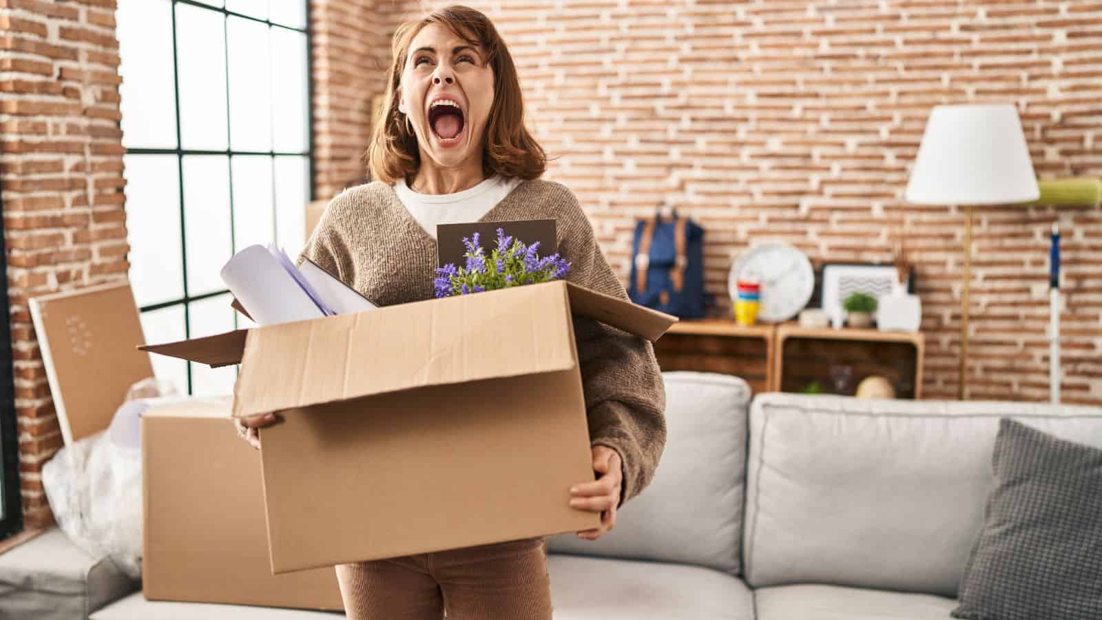 A woman stands in a living room, holding an open cardboard box brimming with items like a small plant and papers. Clad in a brown sweater, she seems to be shouting amidst the brick walls and large window, perhaps about smart hacks for a clutter-free home.