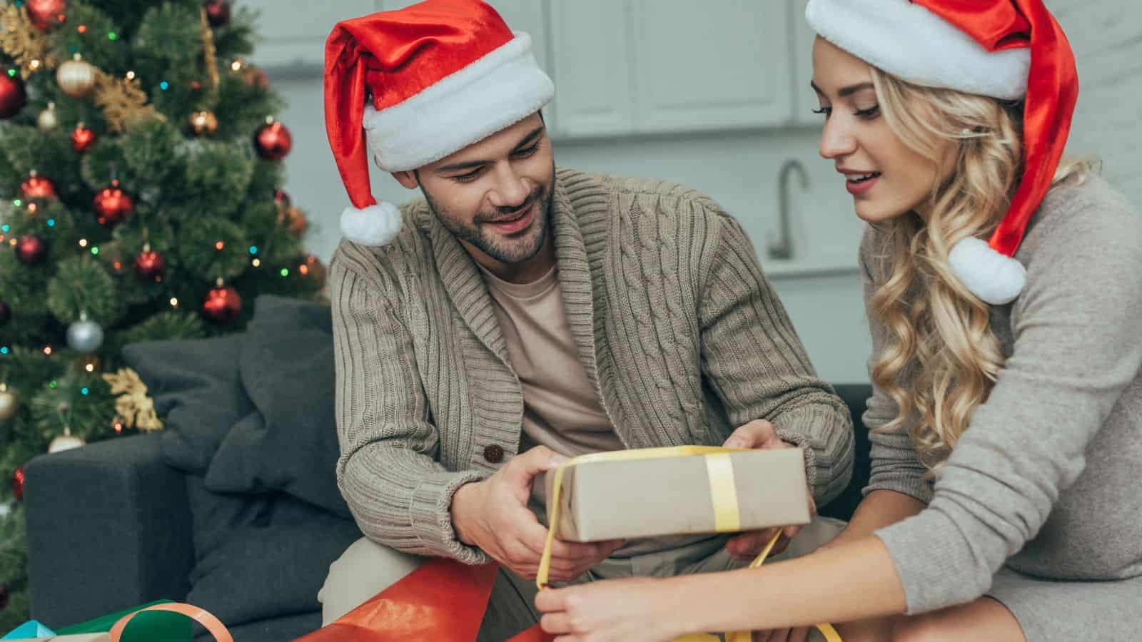 A man and a woman wearing Santa hats are sitting together near a Christmas tree. They are wrapping a gift with brown paper and a yellow ribbon. Various rolls of wrapping paper are scattered around them.