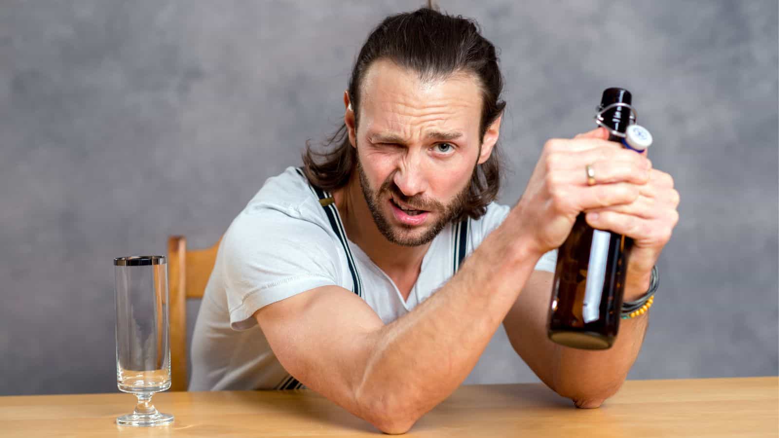 A man with long hair sits at a table, clutching a brown bottle with both hands, his face scrunched in contemplation. An empty glass lurks beside him, hinting at hidden drinking patterns against the plain gray wall.