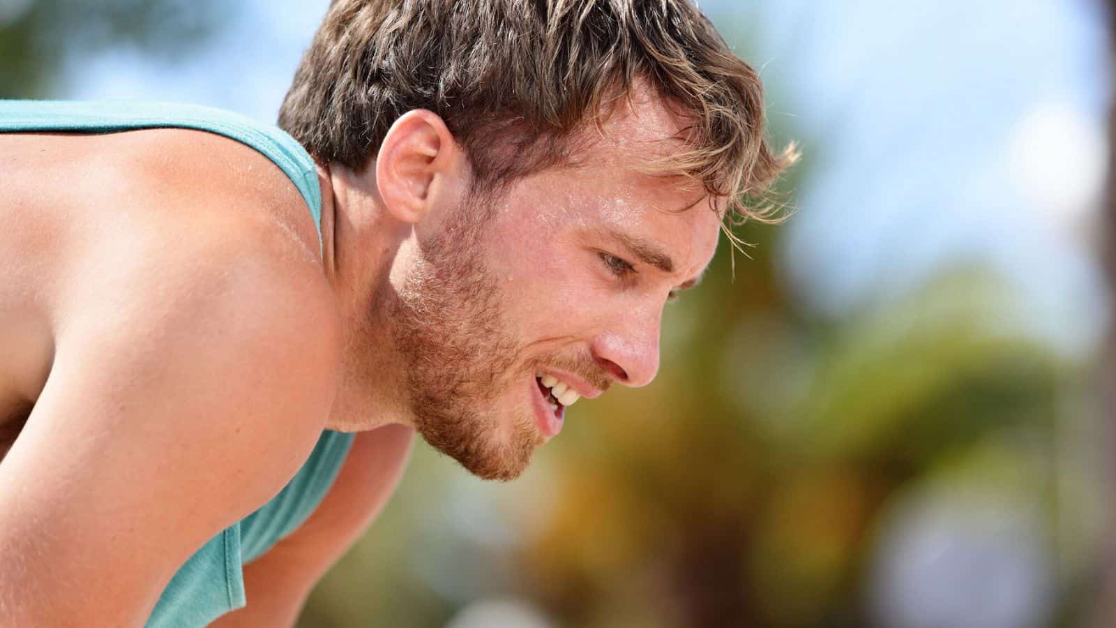 A man with short hair in a turquoise tank top is bent over, appearing out of breath. The background shows blurred greenery and a clear blue sky.