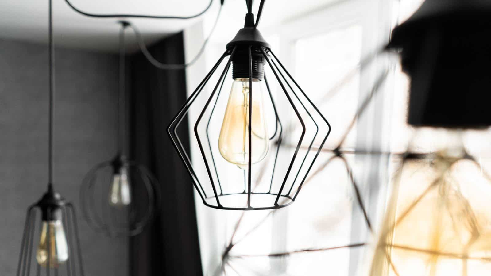 Close-up of modern pendant lights with exposed bulbs and geometric wireframe designs, hanging in a bright room with a large window in the background. The focus is on the central light fixture, highlighting its intricate design.