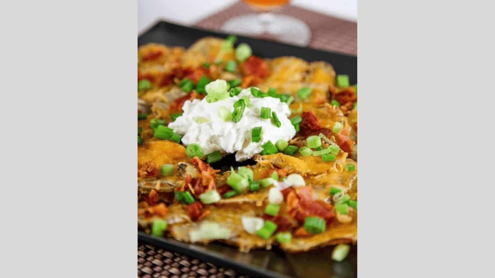 A plate of loaded nachos topped with melted cheese, bacon bits, chopped green onions, and a dollop of sour cream. A drink is blurred in the background.