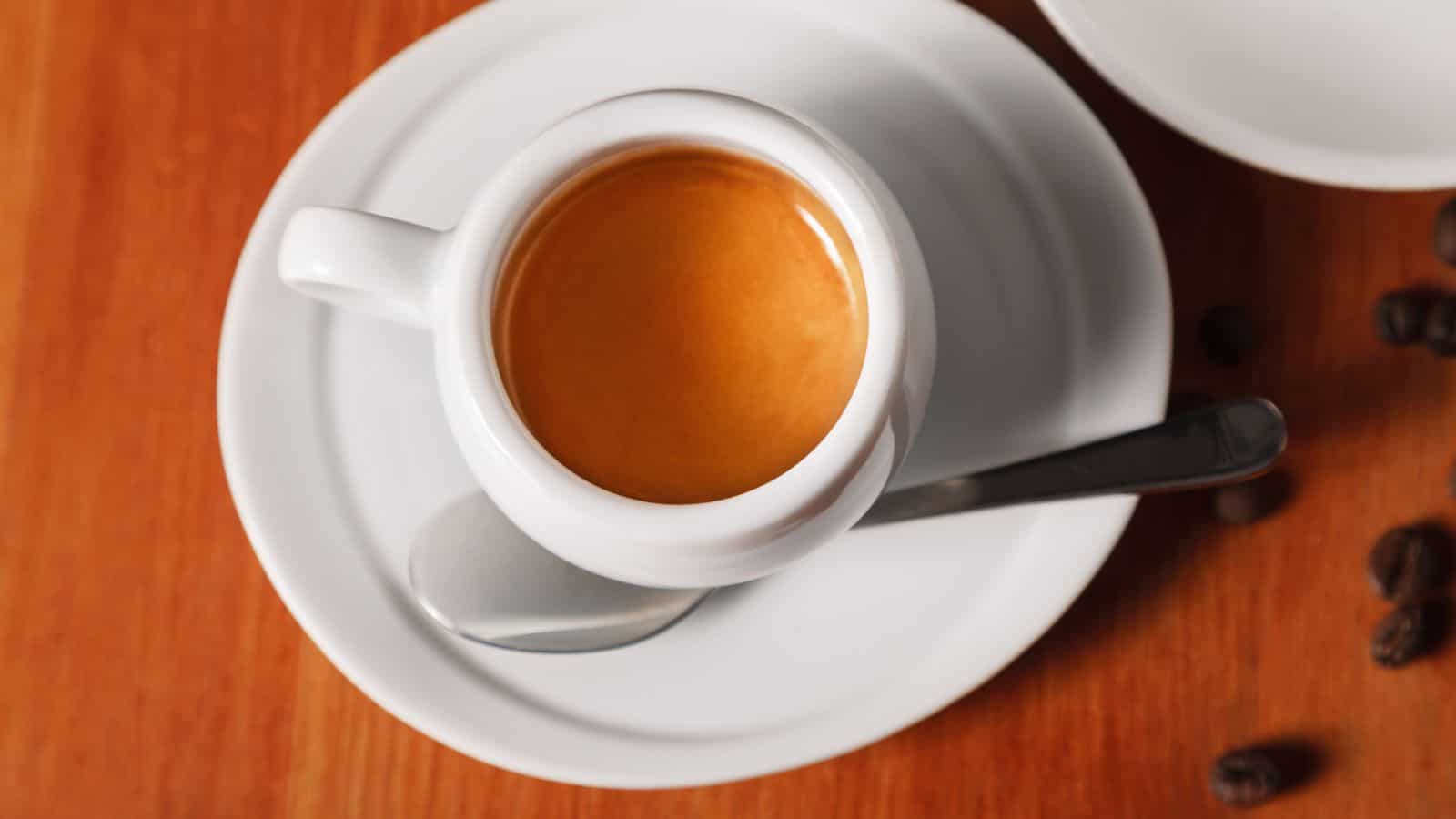 A white cup filled with espresso sits on a matching saucer. A silver spoon is placed on the saucer. The background is a wooden surface with a few scattered coffee beans.