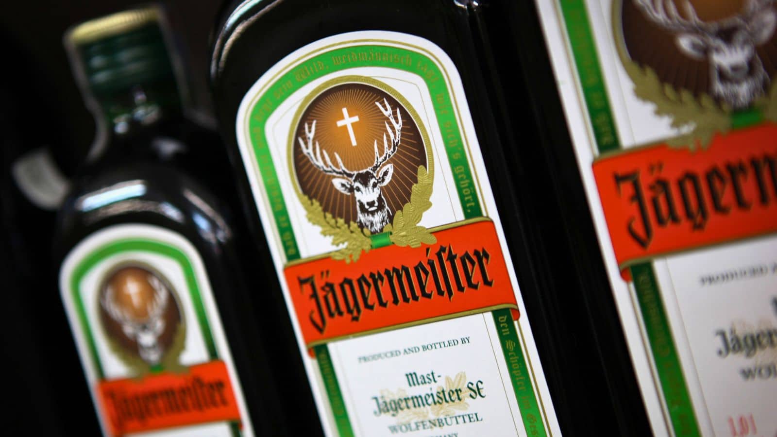 Close-up of J&auml;germeister bottles with a green and orange label featuring a stag with a glowing cross between its antlers. The brand name "J&auml;germeister" is prominently displayed in black and orange lettering.