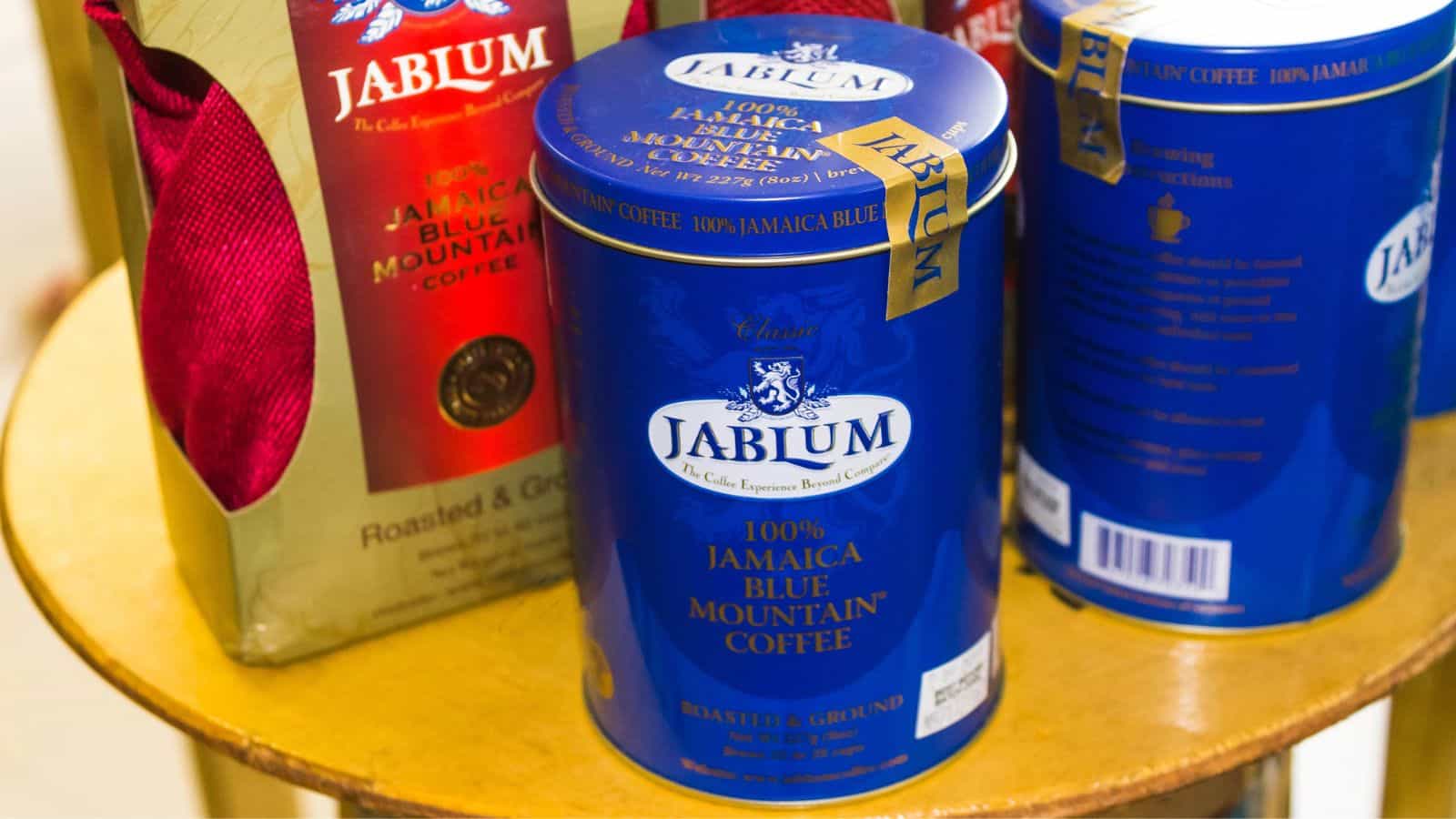 Cans and bags of Jablum 100% Jamaica Blue Mountain coffee are displayed on a wooden table. The packaging is blue with gold accents, featuring the brand name and product details prominently.