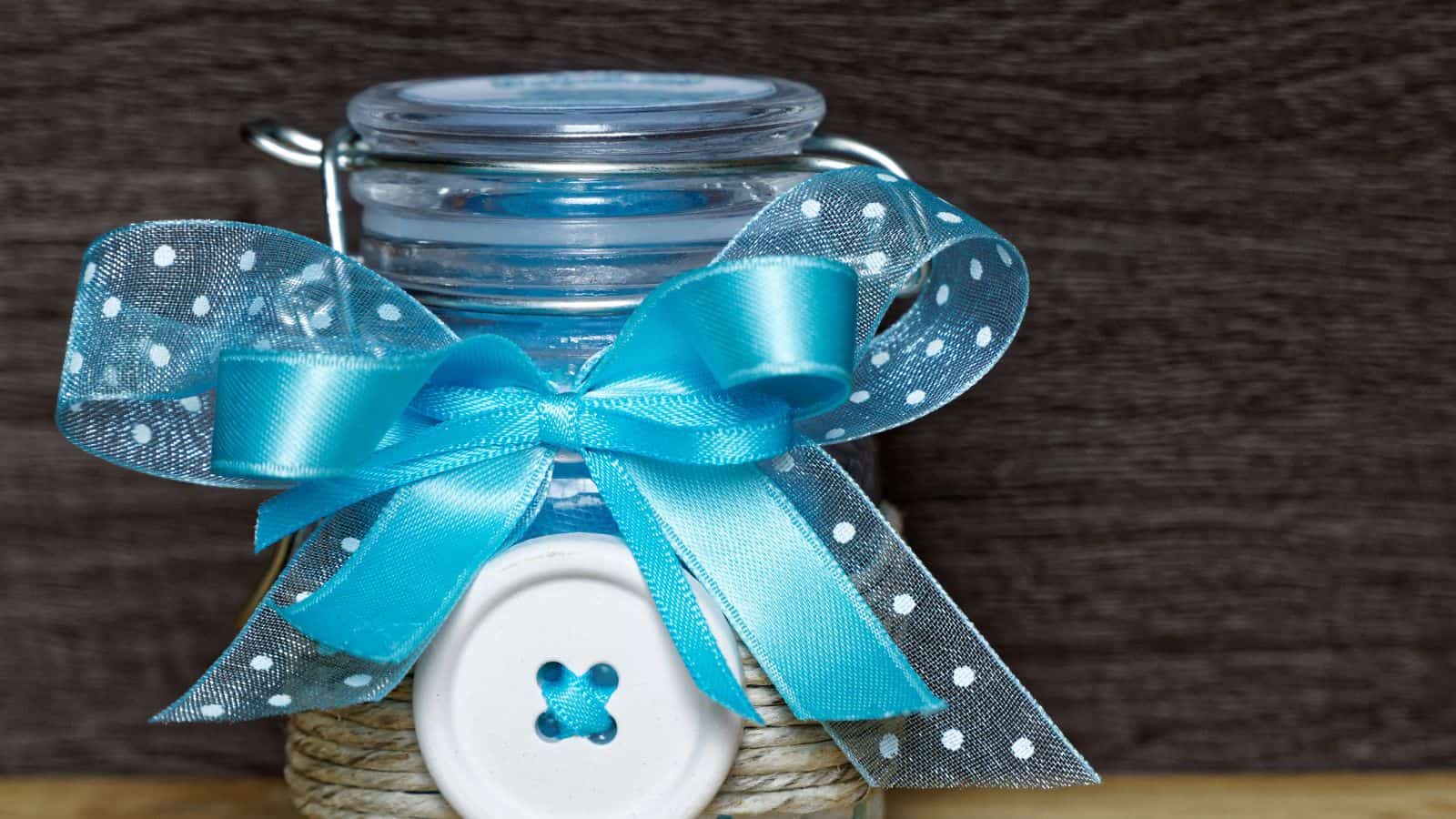 A small glass jar decorated with a blue ribbon tied into a bow, featuring white polka dots. The jar has a white button attached with string around its base. It is set against a dark textured background.