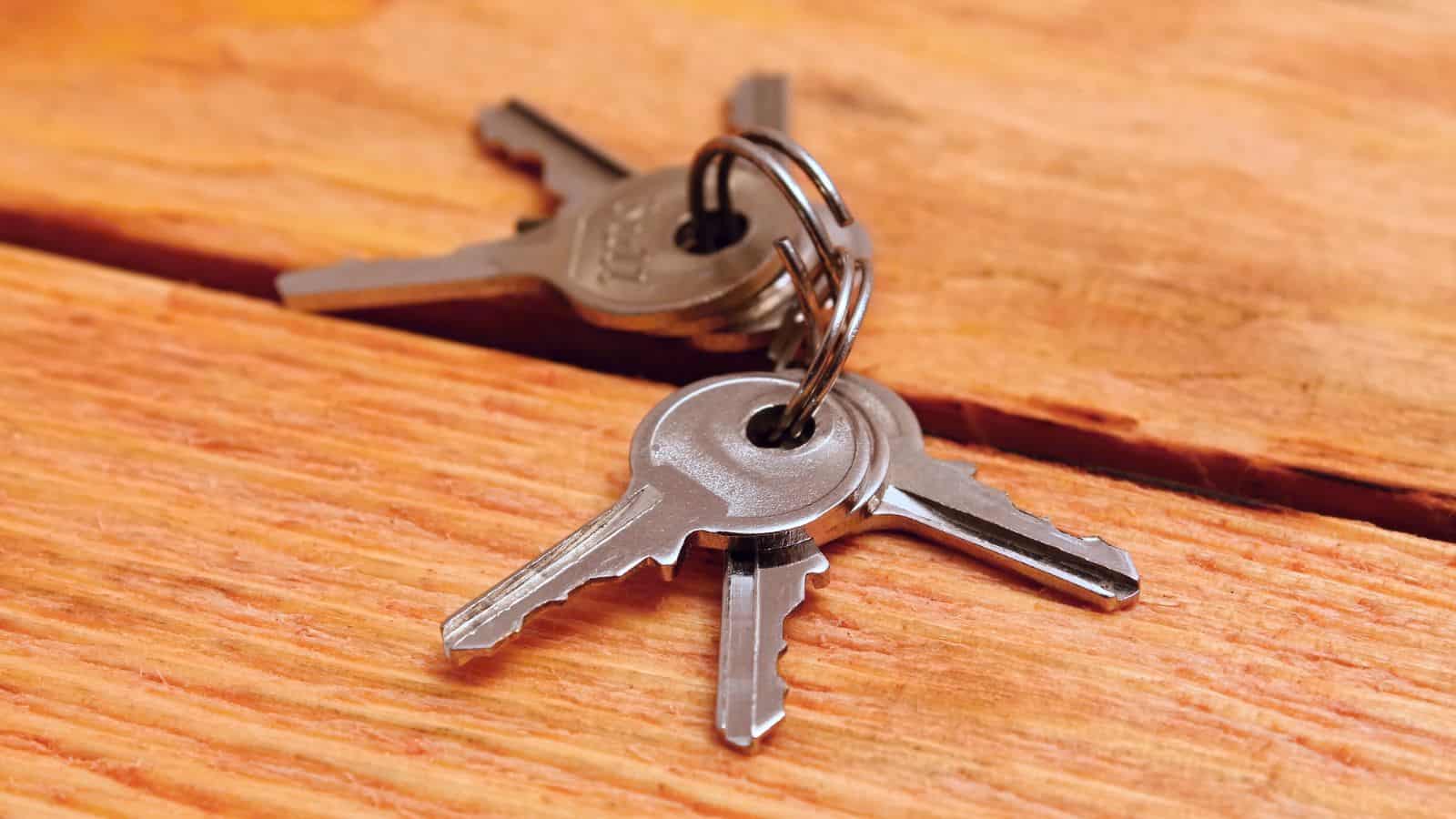 A set of four silver keys on a metal keyring lies on a wooden surface with visible grain patterns. The keys are slightly overlapping each other.