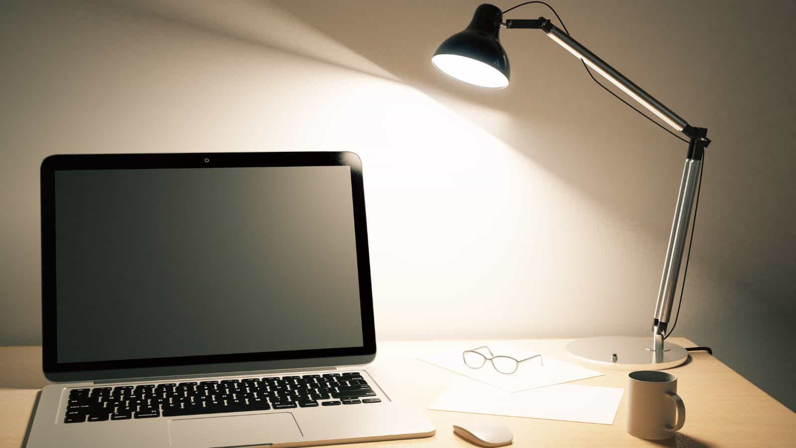 A desk with a laptop displaying a blank screen, illuminated by a modern desk lamp. Nearby are a pair of eyeglasses, a sheet of paper, and a small cup, all on a light-colored wooden surface.