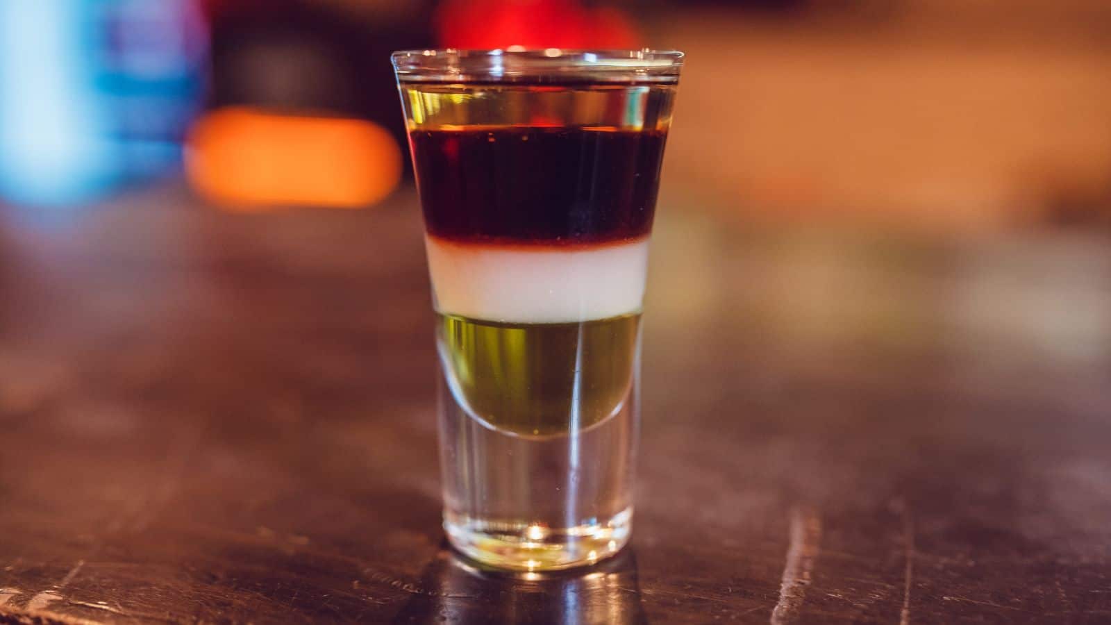 A layered shot glass drink with three distinct layers: a dark brown top layer, a white middle layer, and a green bottom layer. The glass is placed on a reflective wooden surface with a blurred background.