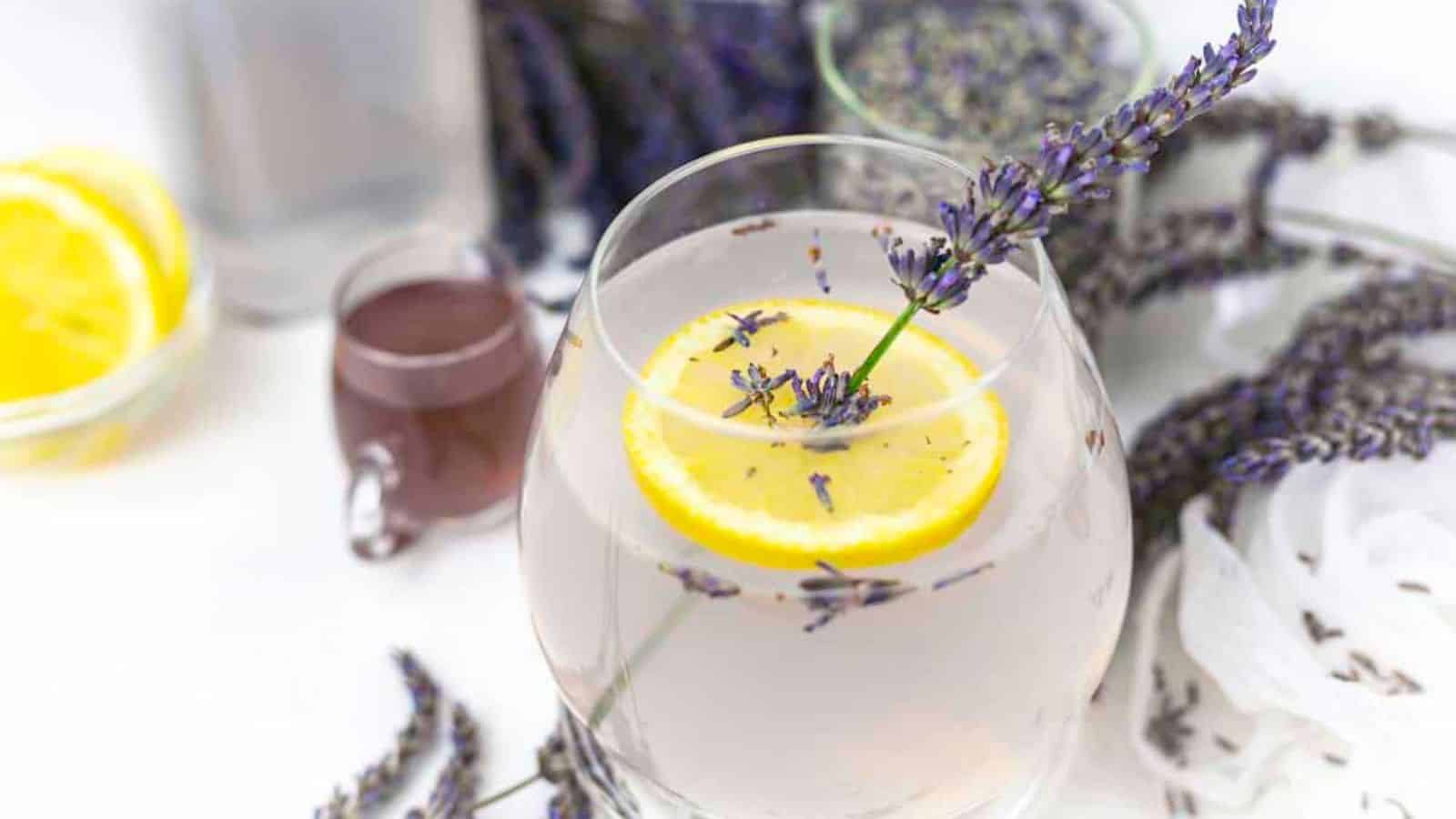 A glass of lavender lemonade with a lemon slice and lavender sprig. There are additional lavender flowers, a small glass of purple liquid, and sliced lemons on a table in the background.