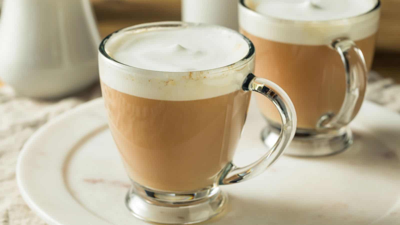 Two clear glass mugs filled with frothy latte are placed on a white plate. The lattes have a creamy foam layer on top. The background is softly blurred, featuring a white pitcher and a light setting.