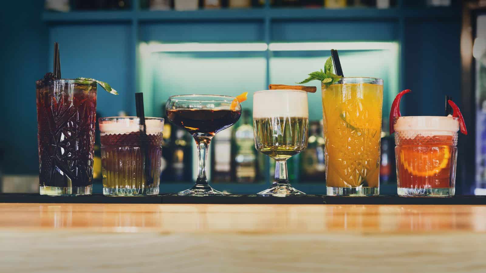 A row of seven colorful cocktails on a bar counter, each in a different style of glass. The drinks vary in color, garnished with items like fruit slices, herbs, and straws. The background shows shelves with blurred bottles.