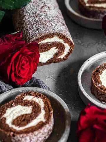 A chocolate Swiss roll with a cream filling is displayed on a grey surface. Slices are neatly arranged on small plates. Red roses and a dark cloth are artfully placed around the dessert.