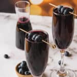 Two glasses filled with a dark purple beverage are garnished with blueberries on skewers. A carafe with the same beverage and a small bowl of blueberries are in the background, set on a marble surface. Warm, blurred lights are visible in the background.