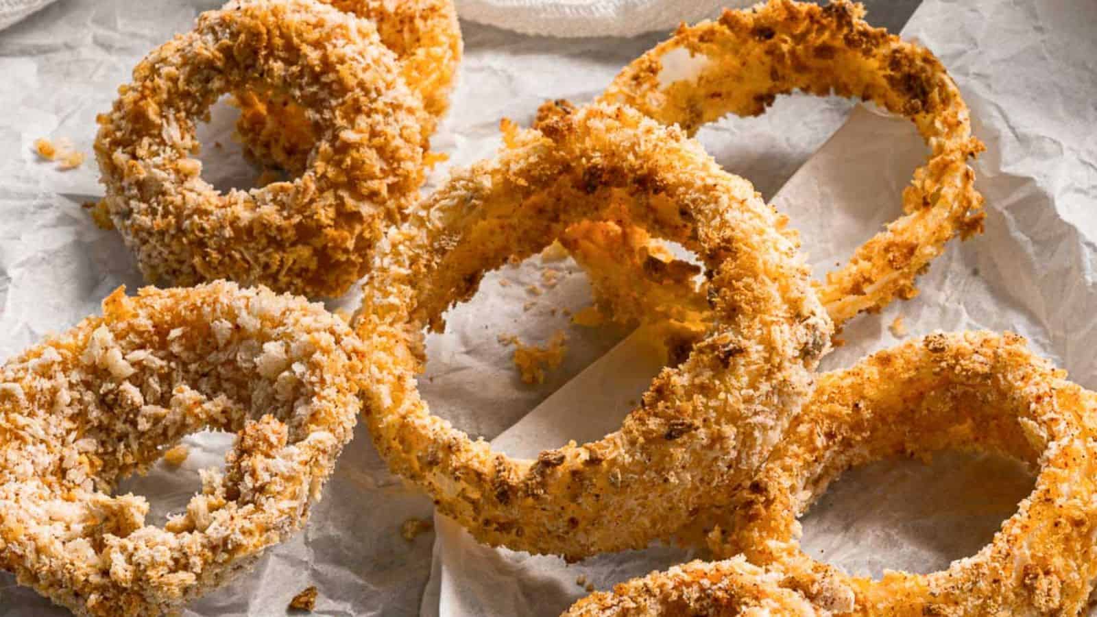 A close-up of several crispy, golden-brown onion rings on crumpled parchment paper. The texture of the breadcrumbs is visible, and a few crumbs are scattered around.