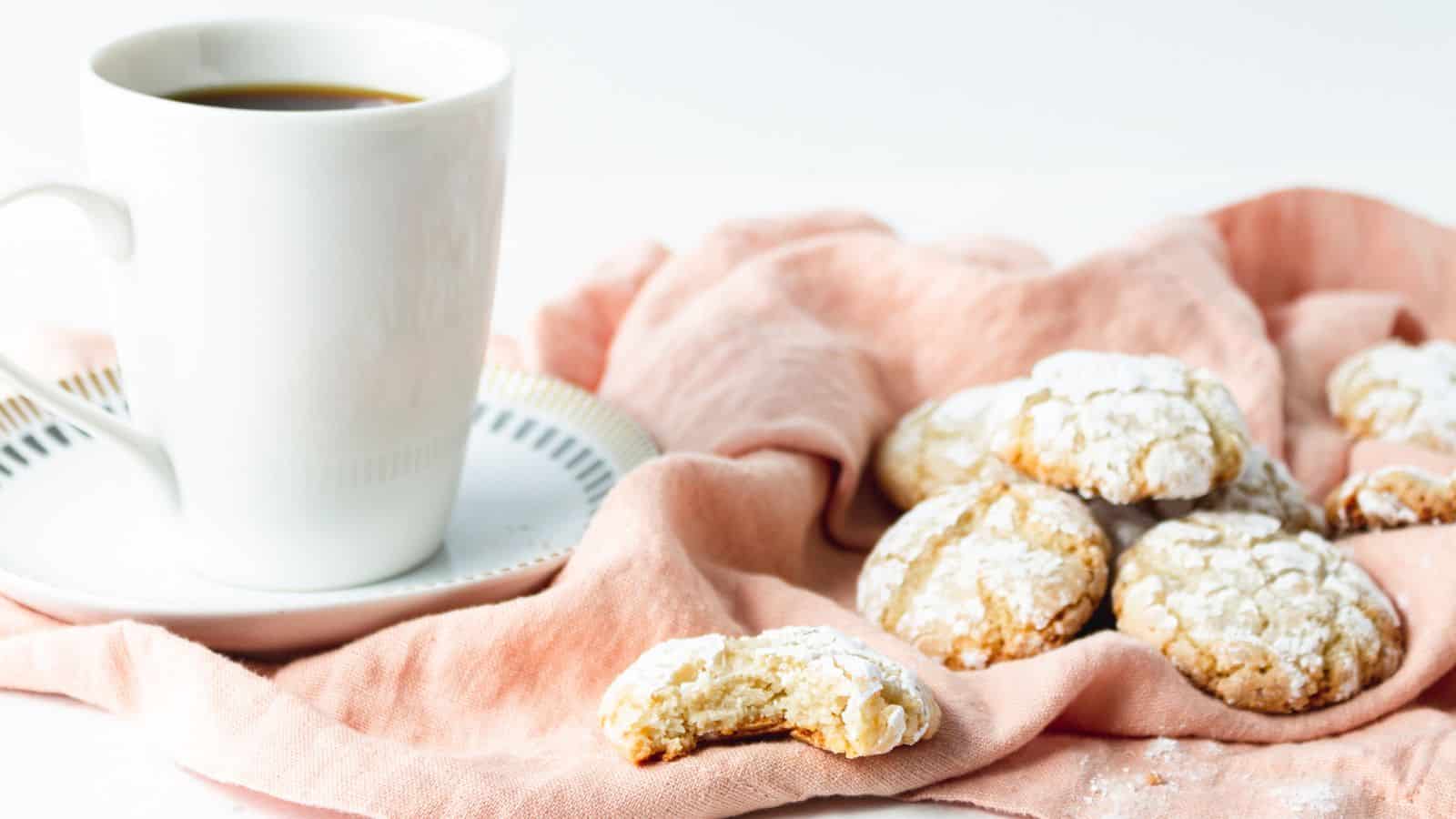 A white cup of coffee sits on a saucer next to a group of powdered cookies on a pink cloth. One cookie has a bite taken out of it. The background is a light, neutral color.