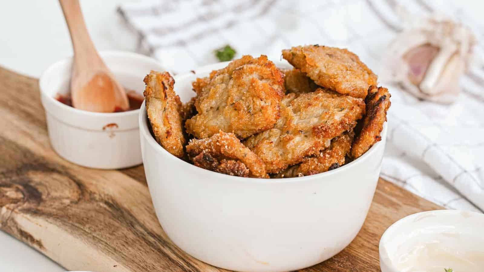 A white bowl filled with crispy, golden-brown fritters sits on a wooden board. A ramekin of dipping sauce with a small wooden spoon is beside it. A striped cloth and a garlic bulb are in the background.