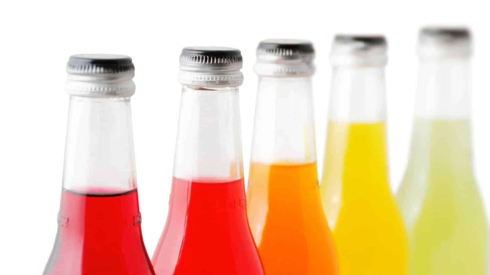 Five glass bottles of soda with metal caps are aligned in a row. The sodas are in different colors: red, orange, yellow, and light green, indicating various flavors. The focus is on the necks and tops of the bottles against a white background.