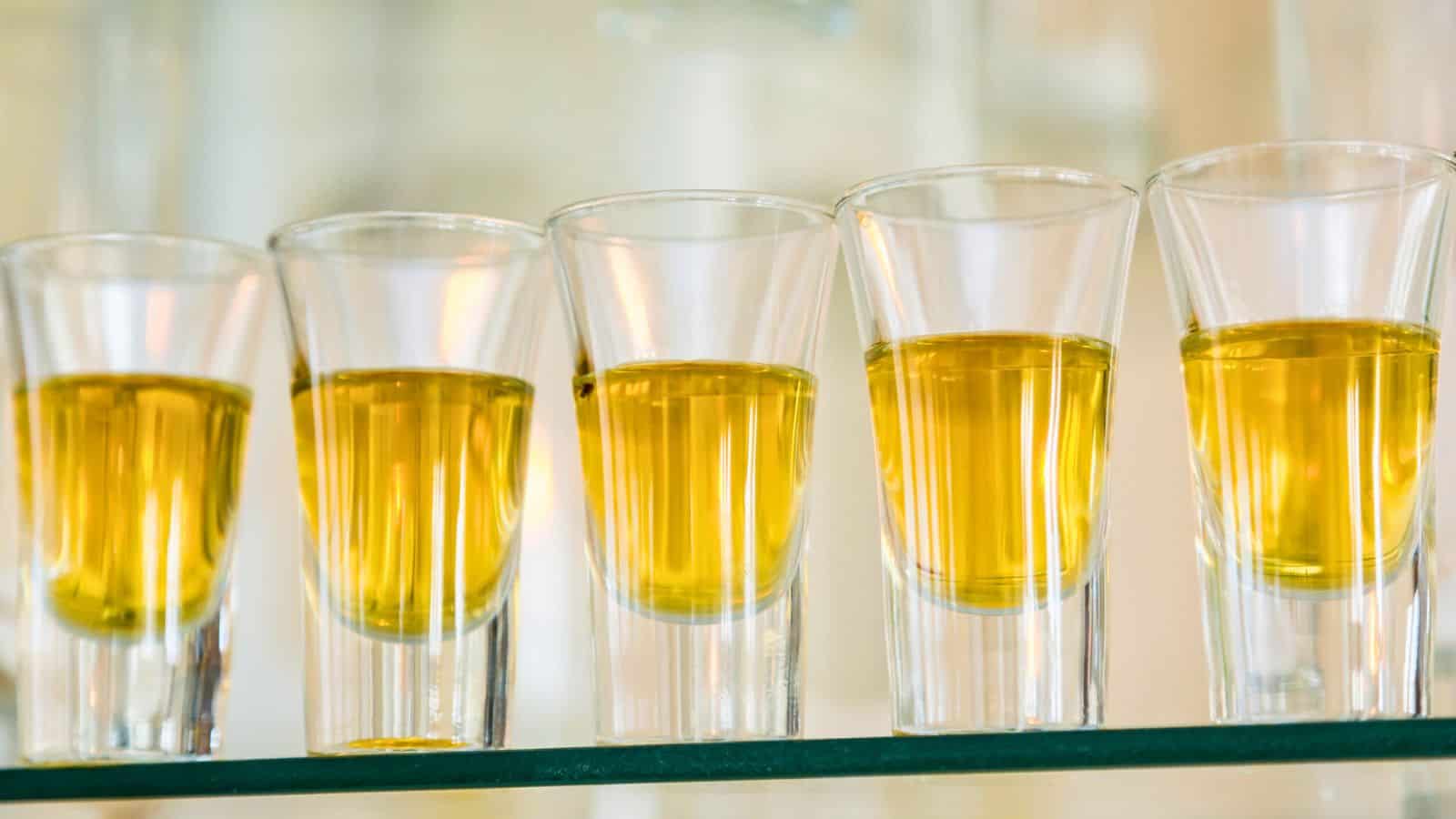 Five shot glasses filled with a golden liquid are lined up side by side on a glass surface. The background is softly blurred, highlighting the glasses.