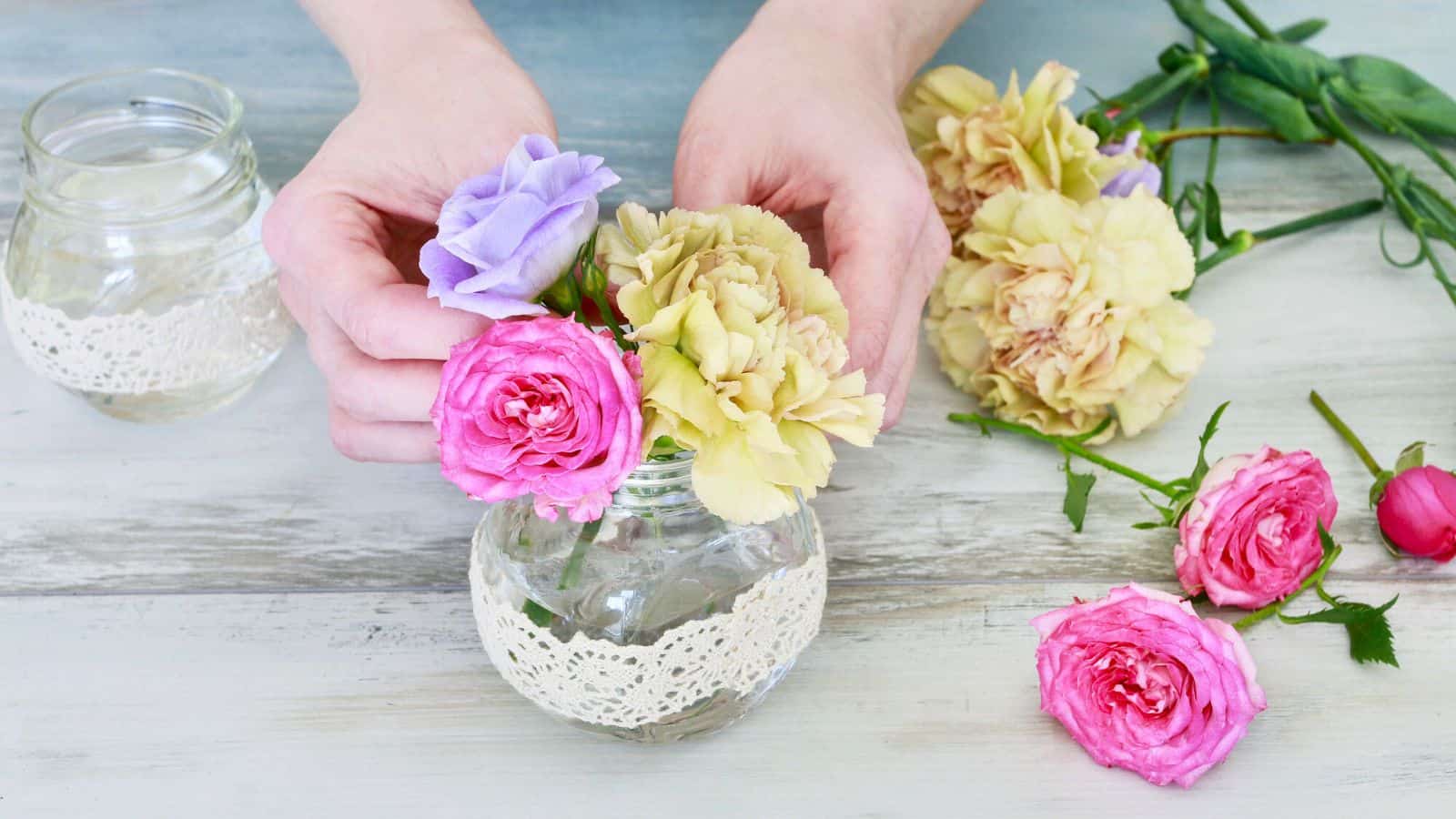 Hands arranging pink, yellow, and purple flowers in a small glass jar with lace decoration, placed on a light wooden surface. Additional flowers and a jar are nearby.