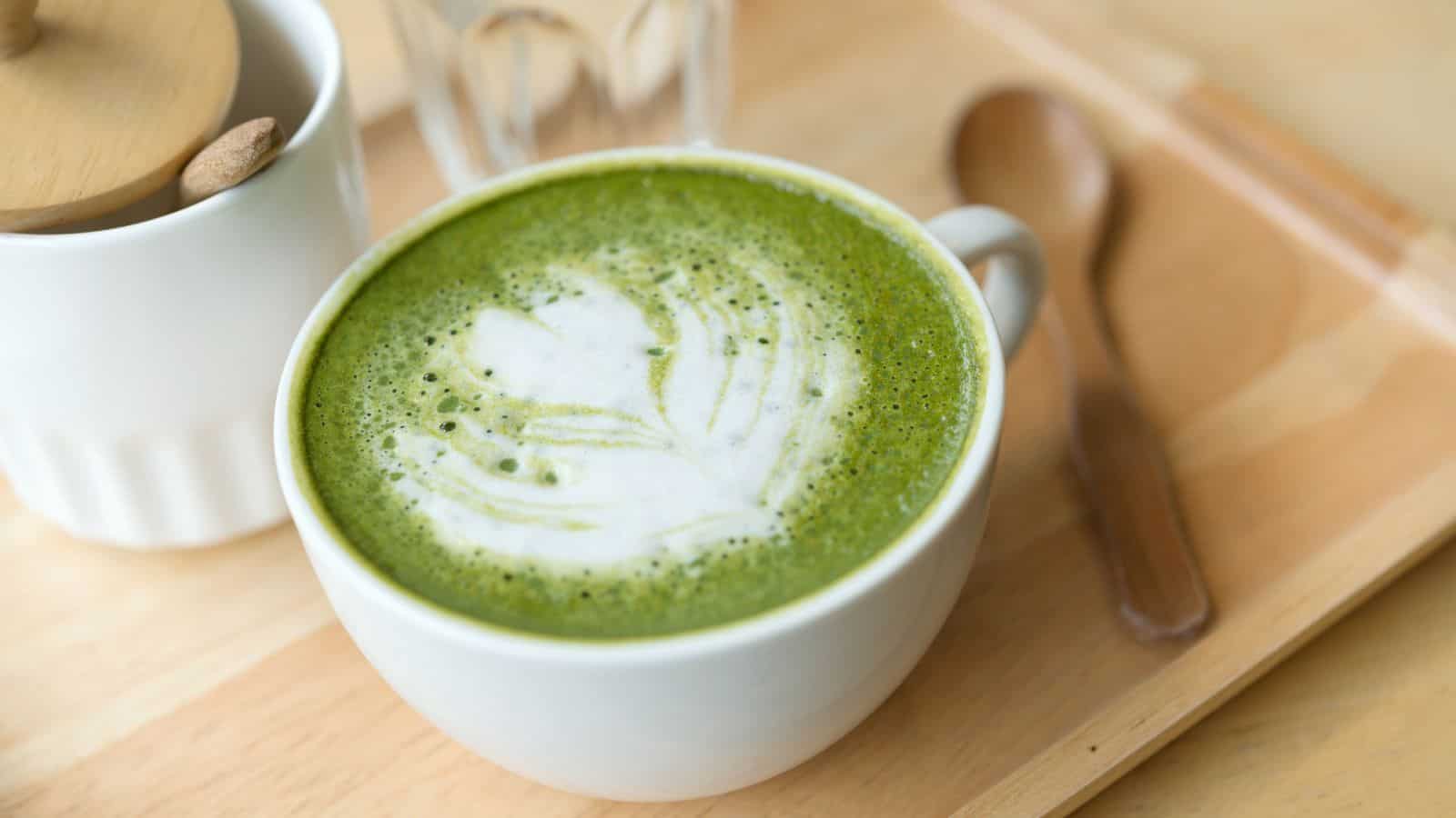 A white cup filled with a matcha latte, featuring intricate latte art on the frothy green surface. The cup is placed on a wooden tray beside a wooden spoon and a small lidded container.