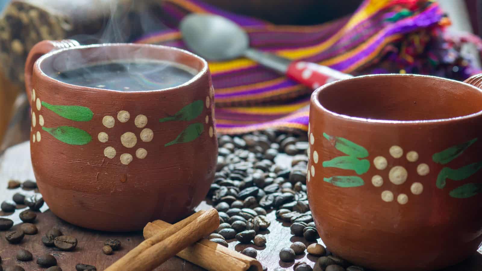 Two clay mugs with painted floral designs filled with black coffee, surrounded by scattered coffee beans and cinnamon sticks. A colorful woven cloth and a spoon are in the background. Steam rises from the hot coffee.