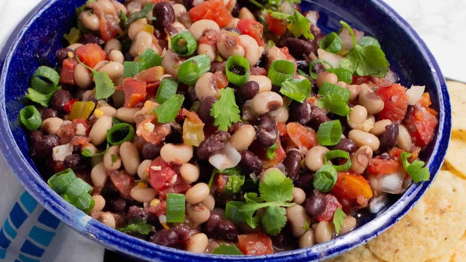 A blue bowl filled with a colorful bean salad, featuring black beans, black-eyed peas, diced tomatoes, chopped green onions, and fresh cilantro. The bowl is placed on a table alongside some tortilla chips.