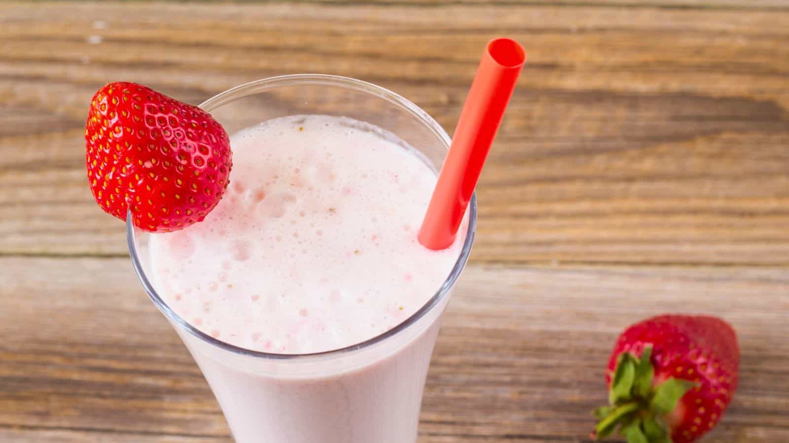 A glass of strawberry milkshake with a red straw and a strawberry garnish sits on a wooden surface. Another strawberry is nearby on the table.