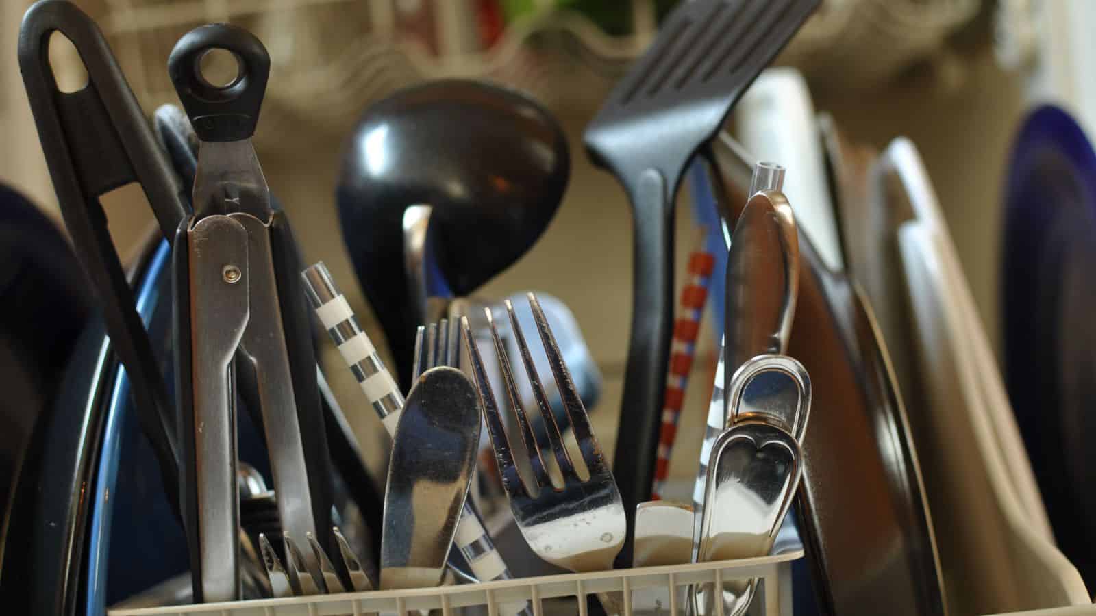A close-up image of assorted kitchen utensils in a dish rack, including forks, knives, a ladle, a spatula, and a can opener. They have various handles, some striped and others plain, with plates in the background.