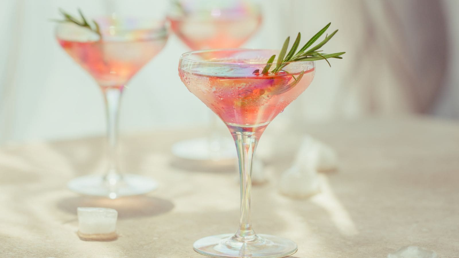 A close-up of three elegant cocktail glasses filled with a pink beverage, each garnished with a sprig of rosemary. The glasses are set on a light-colored surface, with scattered ice cubes in soft focus in the background.
