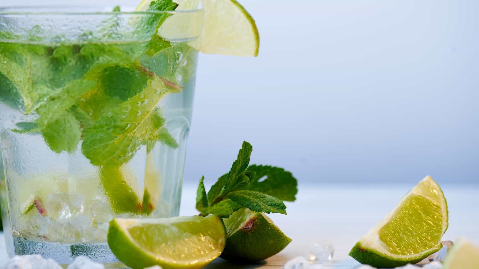 A clear glass filled with a mojito cocktail, featuring mint leaves, lime wedges, and ice cubes. Several lime wedges and mint leaves are scattered around the base of the glass on a light surface. The background is softly blurred.