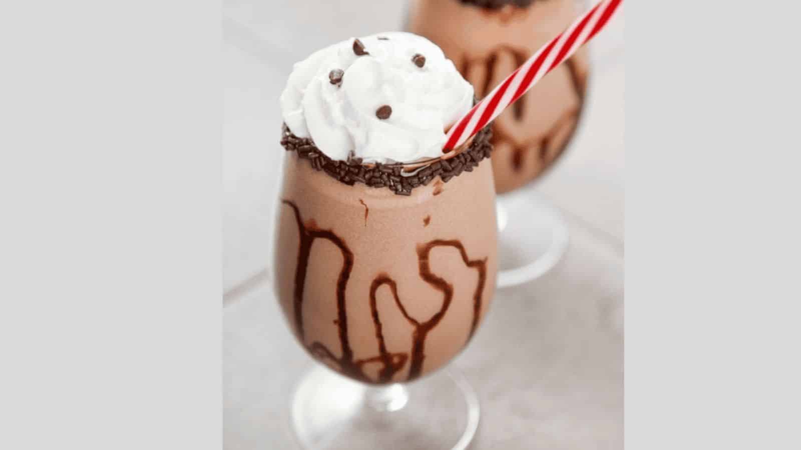 Two chocolate milkshakes with whipped cream are topped with chocolate sprinkles. The glasses are drizzled with chocolate syrup, and each has a red and white striped straw. They are placed on a light-colored surface.