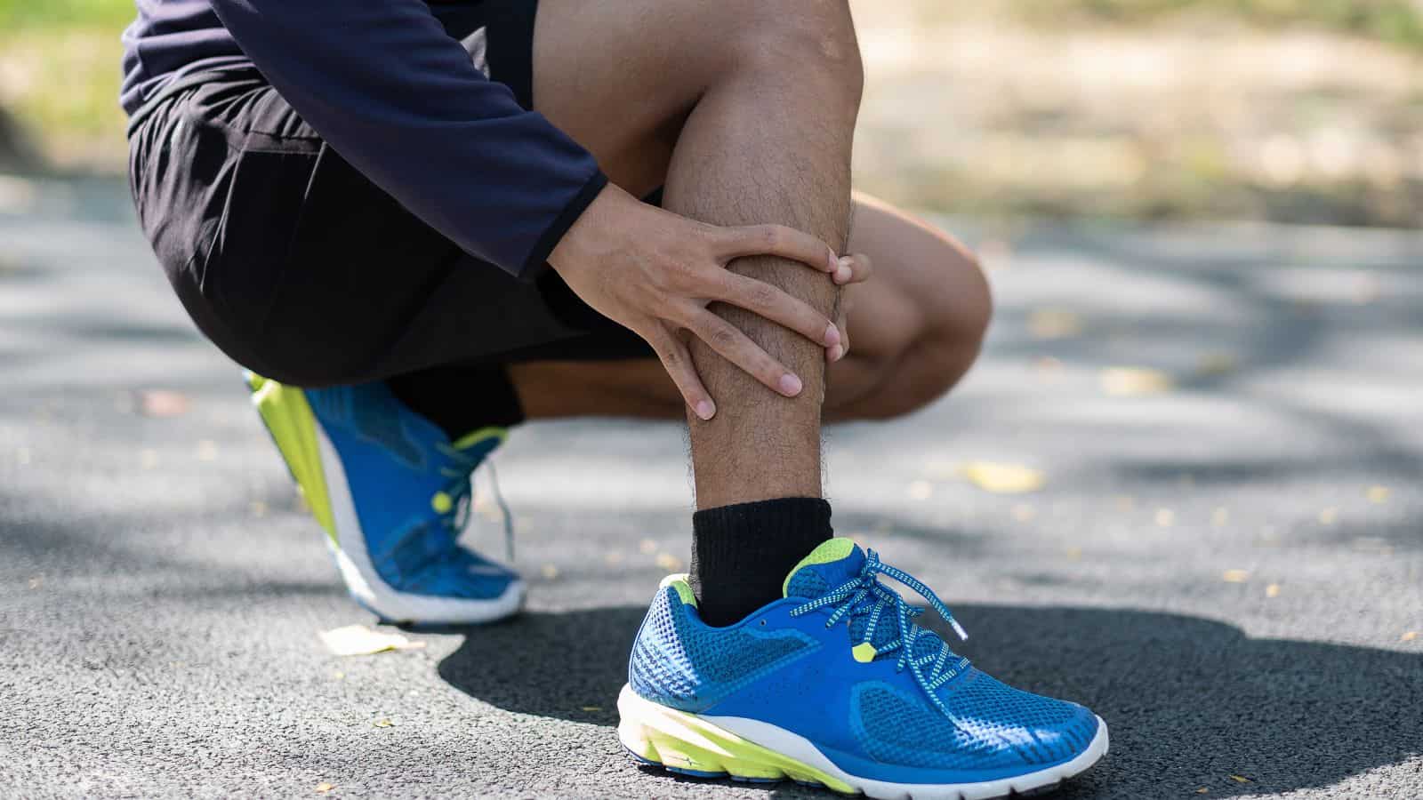 A person in athletic clothing crouches on a paved surface, touching their lower leg as if examining or massaging it. They are wearing blue running shoes with bright yellow and green accents. Fallen leaves are scattered on the ground.