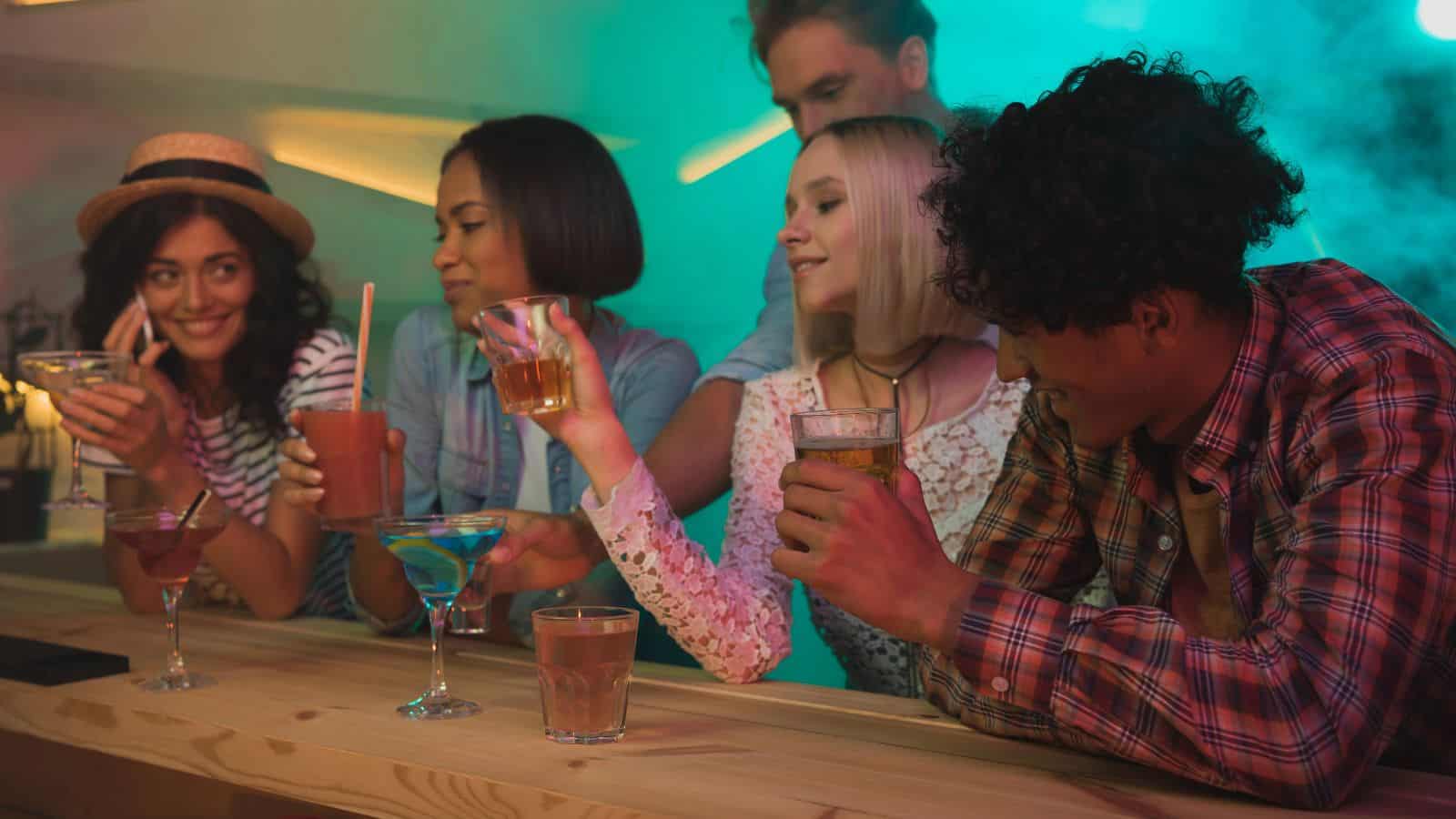 Five people are at a bar holding various drinks. Some are smiling and engaging in conversation. The atmosphere is lively, with colorful lighting and a relaxed ambiance.