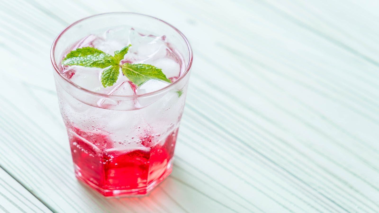A clear glass filled with a red, fizzy drink is topped with ice cubes and a sprig of mint. The glass is placed on a light-colored wooden surface.