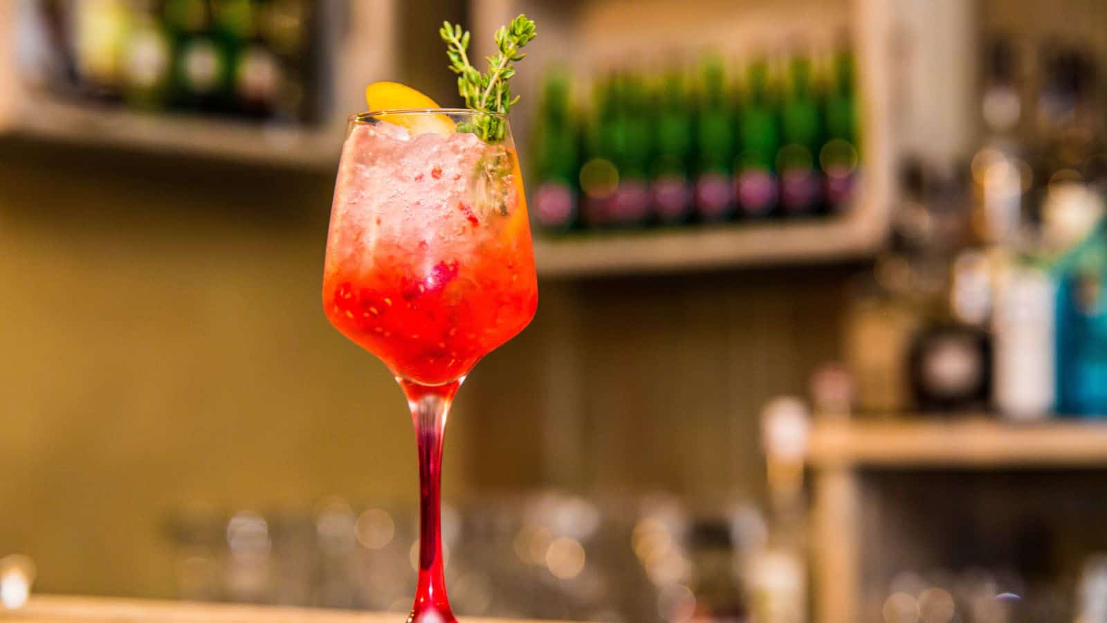 A tall glass filled with a red cocktail sits on a bar counter. The drink is garnished with a sprig of greenery and a small orange slice. The background is blurred, showing shelves with bottles.