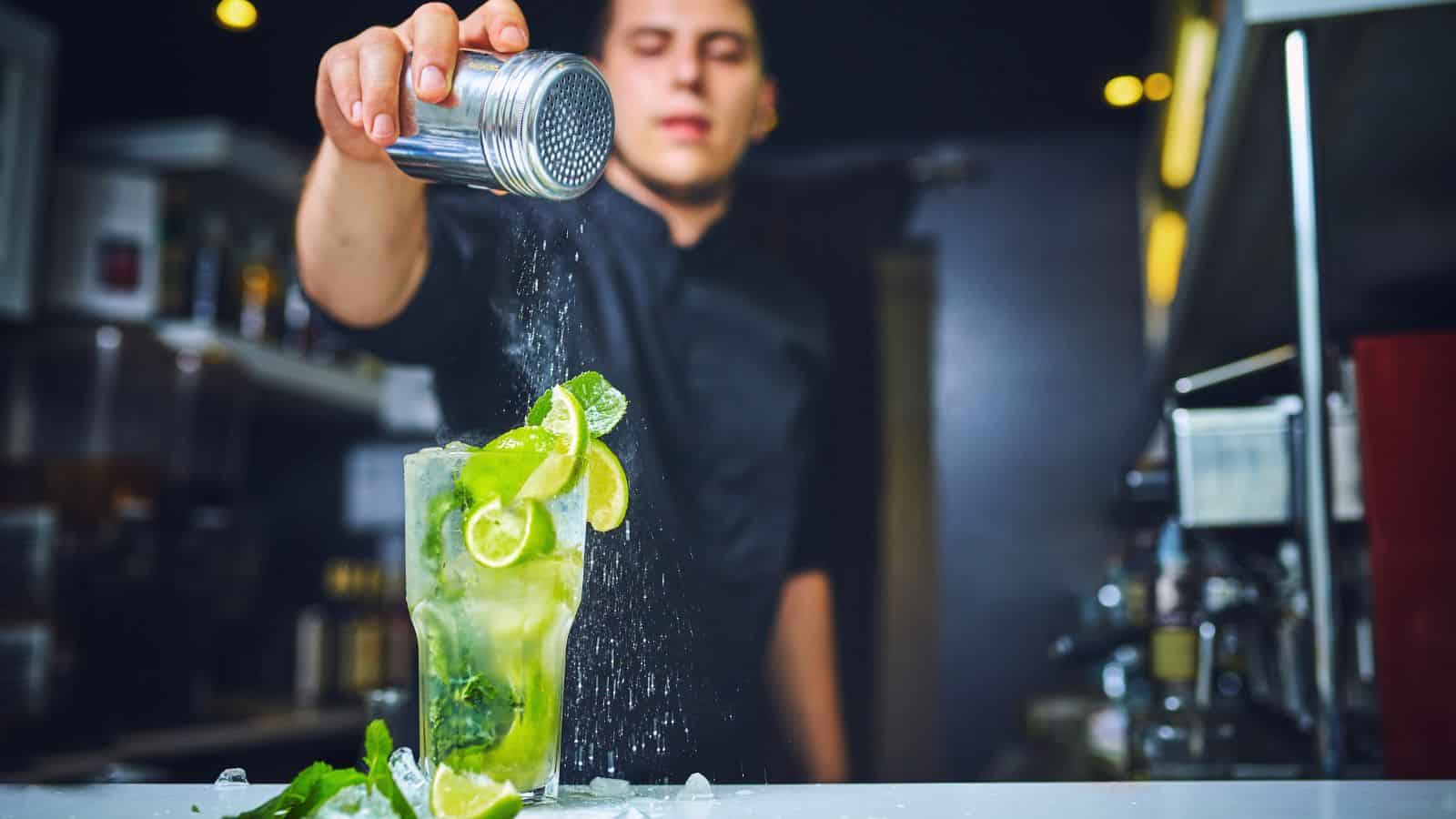 A bartender in a black shirt sprinkles sugar over a cocktail in a tall glass. The drink is garnished with lime wedges and mint leaves. The background features bar shelves with various bottles.