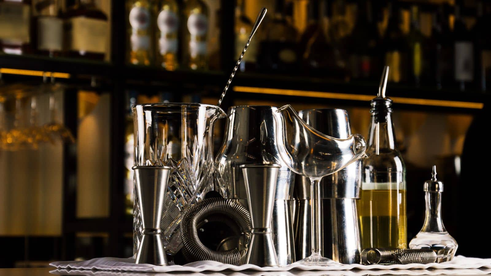 A collection of bar tools including jiggers, a mixing glass, a strainer, and a stirring spoon are arranged on a bar counter. Bottles are visible on shelves in the background, suggesting a bar or cocktail preparation setting.