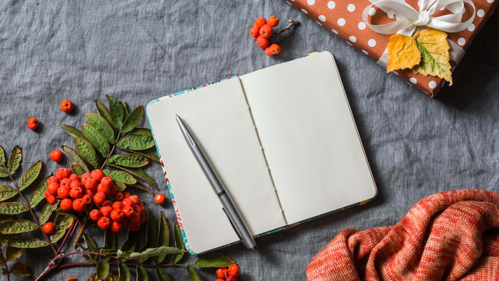 An open blank notebook with a pen rests on a gray surface. To the left are sprigs of red berries and green leaves. A wrapped gift with an orange bow and a piece of fabric are on the right, adding a cozy autumnal feel.