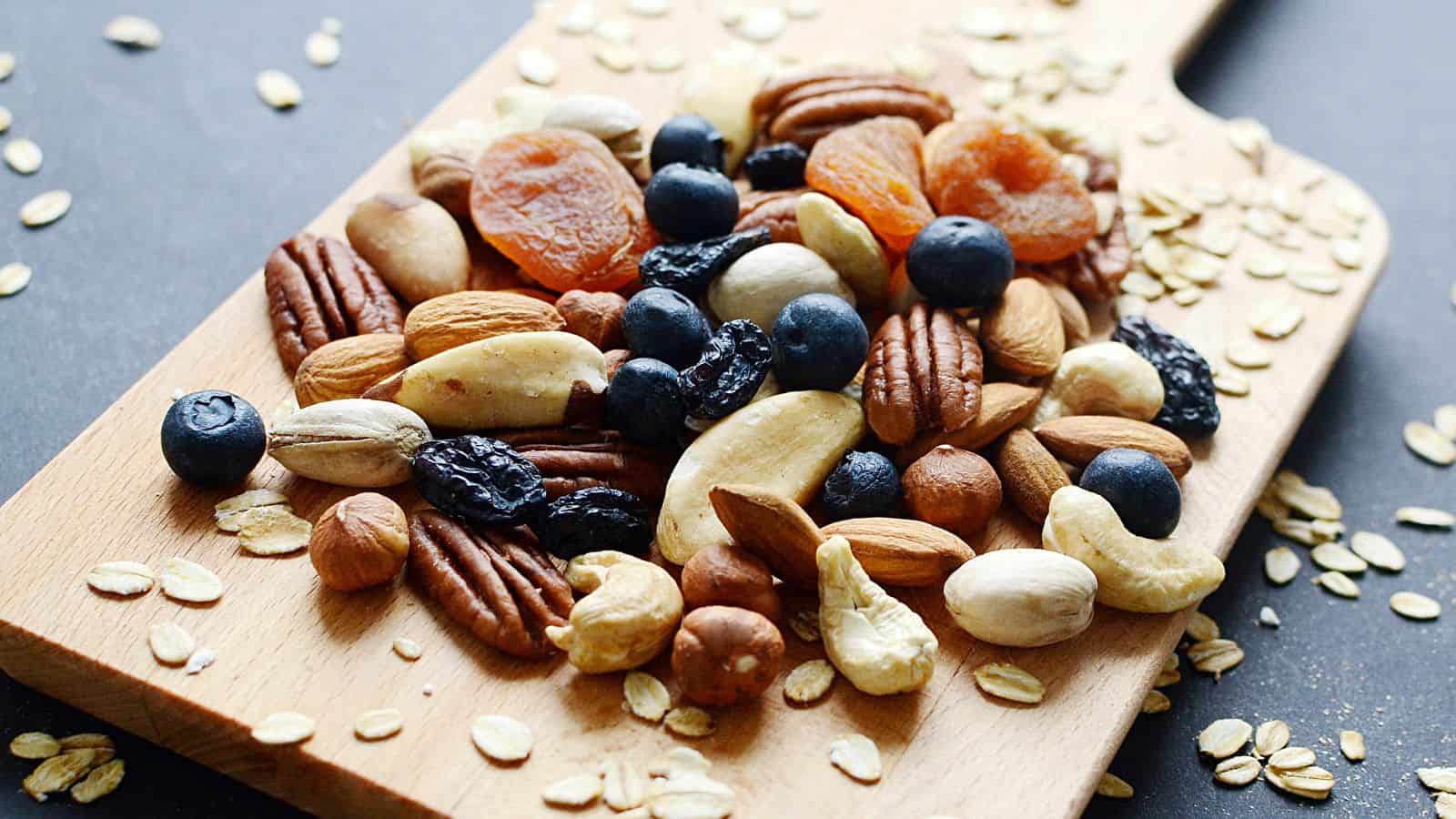 A wooden cutting board with a pile of mixed nuts and dried fruits, including pecans, cashews, almonds, dried apricots, dried blueberries, and oats around the edges. The surface is dark gray.