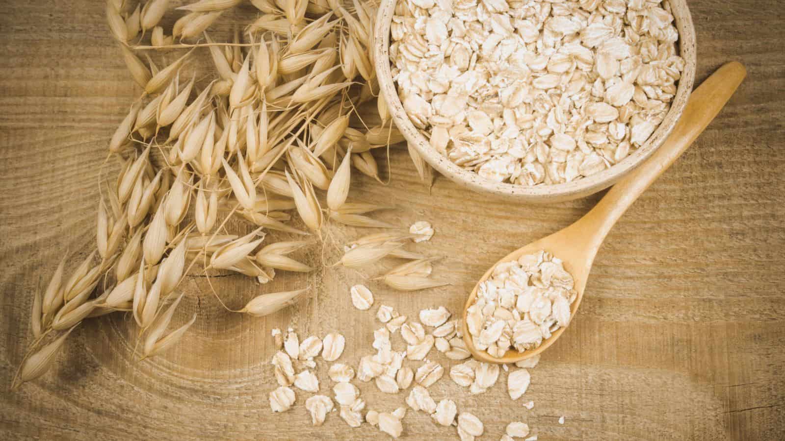 A wooden table with a bowl of rolled oats and a wooden spoon partially filled with oats. A bundle of dry oat stalks is placed beside the bowl, and a few loose oats are scattered on the table.