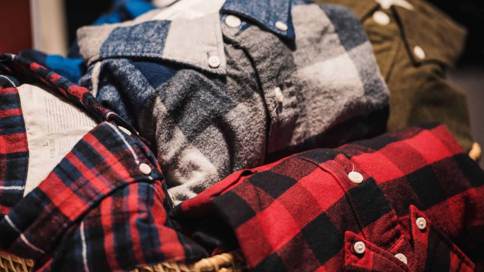 A basket filled with folded flannel shirts in various plaid patterns and colors, including blue, gray, and red. The shirts have button closures and visible collars, and are arranged neatly in the basket.