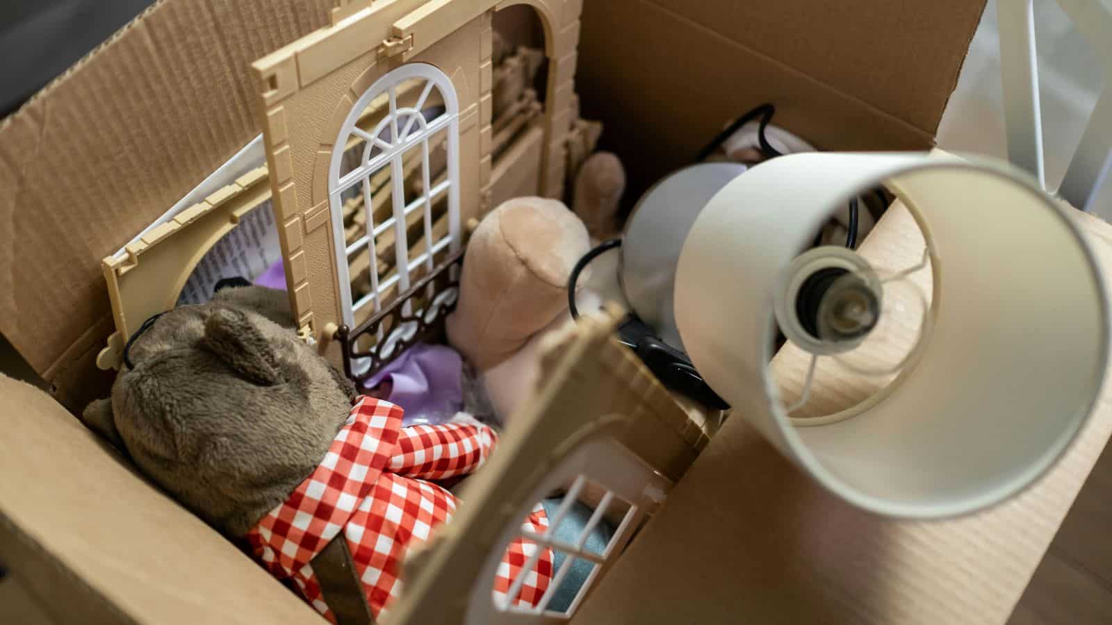 An open cardboard box containing a teddy bear in red checkered clothing, a lampshade, and various plastic pieces resembling parts of a dollhouse. The box appears to be packed for moving or storage.