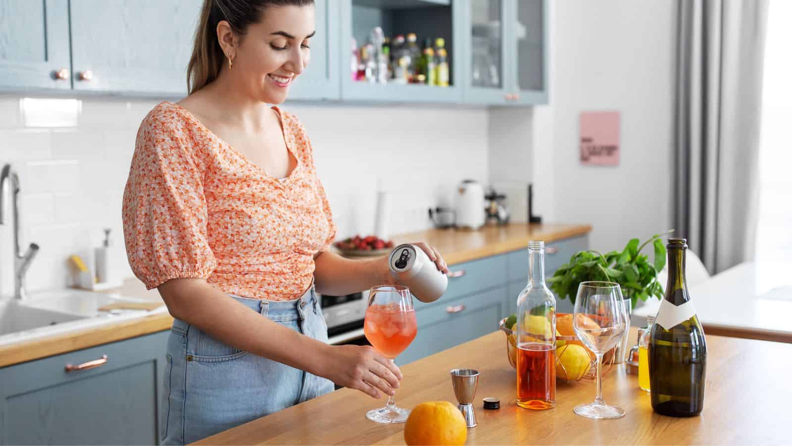 A woman stands in a kitchen pouring a drink into a glass filled with ice cubes. The counter has a bottle of red liquid, wine bottles, a jigger, and citrus fruits. She wears a floral top and smiles while preparing the beverage.