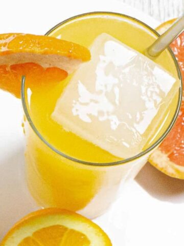 Two glasses of orange-colored drinks with large ice cubes and orange slices on the rims. A grapefruit half is placed beside the drinks on the white surface.