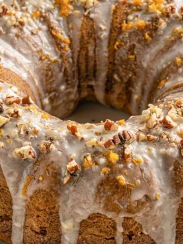 A Bundt cake on a white plate is topped with a thick white glaze and sprinkled with crushed nuts. The cake has a golden-brown color and is set on a light-colored surface.