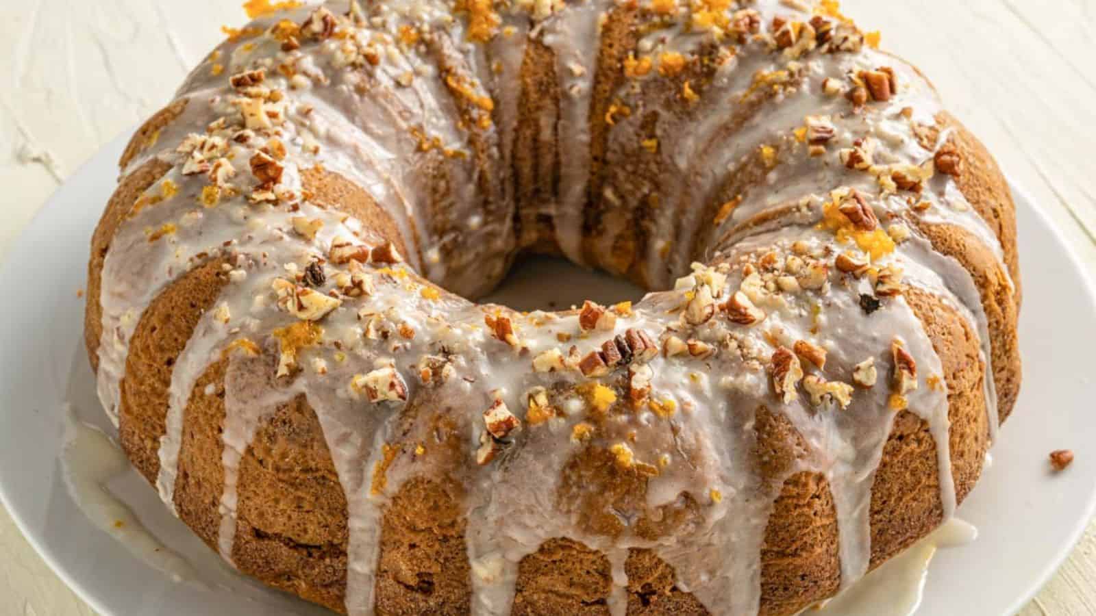 A Bundt cake on a white plate is topped with a thick white glaze and sprinkled with crushed nuts. The cake has a golden-brown color and is set on a light-colored surface.