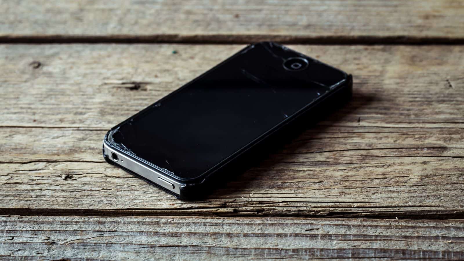 A smartphone with a cracked screen lies on a rustic wooden surface. The phone appears off, and its casing is worn, suggesting frequent use. The wooden background features visible grain and texture.