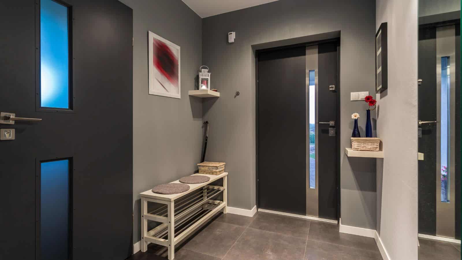 A modern hallway with dark doors and tile flooring. There's a small shelf with cushions, a hanging red and white abstract painting, a wicker basket with red flowers on the right, and a folded umbrella near the corner.