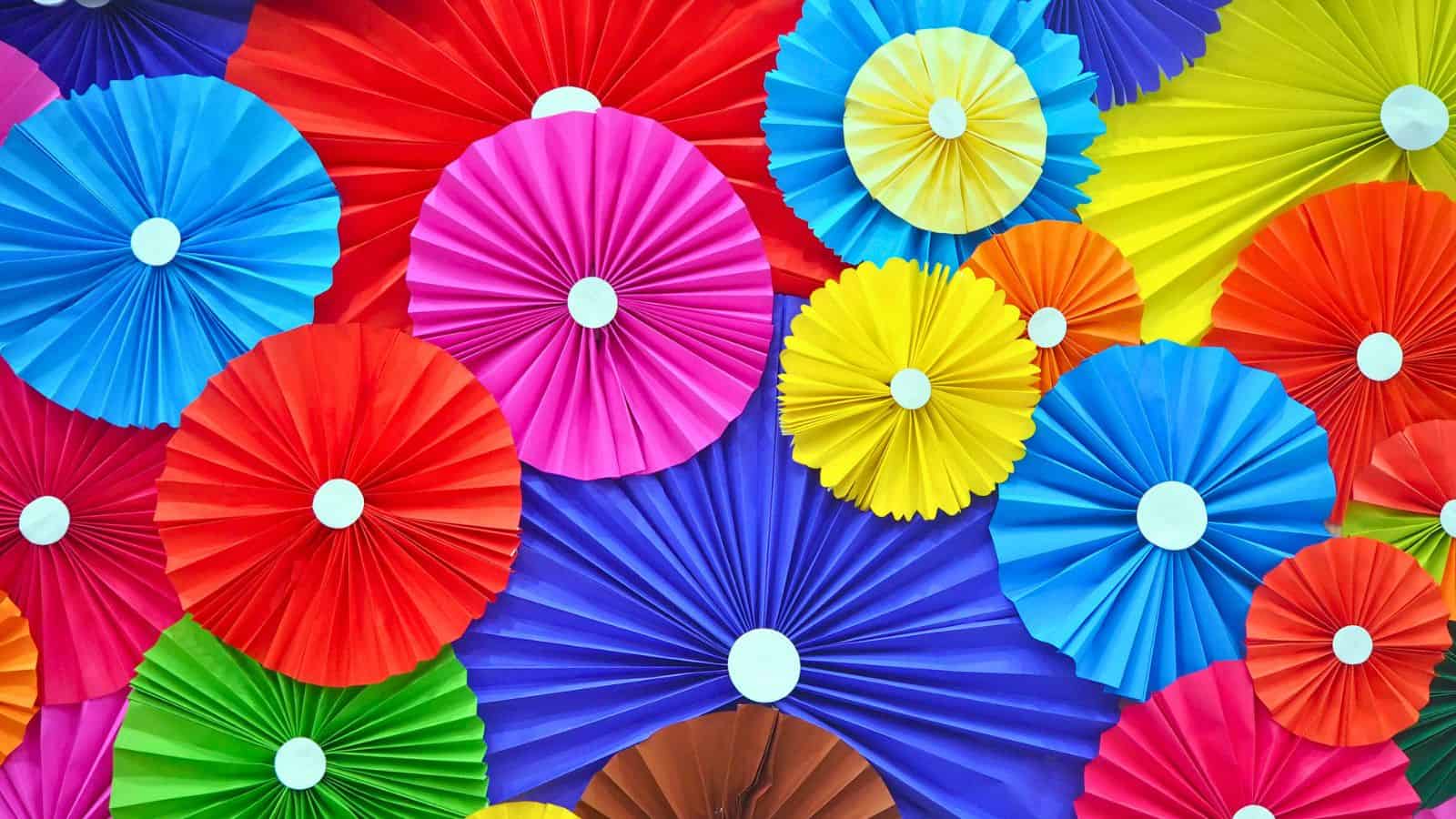 A colorful arrangement of folded paper pinwheels in various sizes, featuring vibrant shades of red, blue, pink, yellow, green, purple, and orange, with white centers. The pinwheels overlap, creating a dynamic and festive pattern.